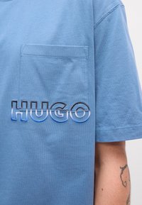 Modrá bavlnená tričko s vreckom na hrudi a logom "HUGO" v prechádzajúcich farbách. Krátke rukávy a voľný strih s viditeľným šitím.