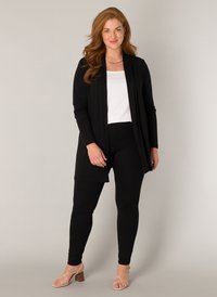 Zwarte open cardigan over een witte top, gecombineerd met zwarte leggings en tan strappy heels. Glad textuur met een relaxte pasvorm.