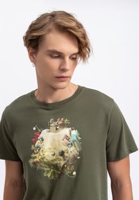T-shirt in cotone verde oliva con una stampa grafica dettagliata di un paesaggio fantasioso con vari oggetti e figure in colori vivaci.