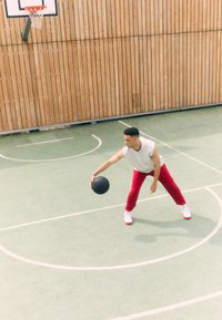 A male figure in a sleeveless gray shirt and red pants dribbles a black basketball on a green court with a wooden backdrop.