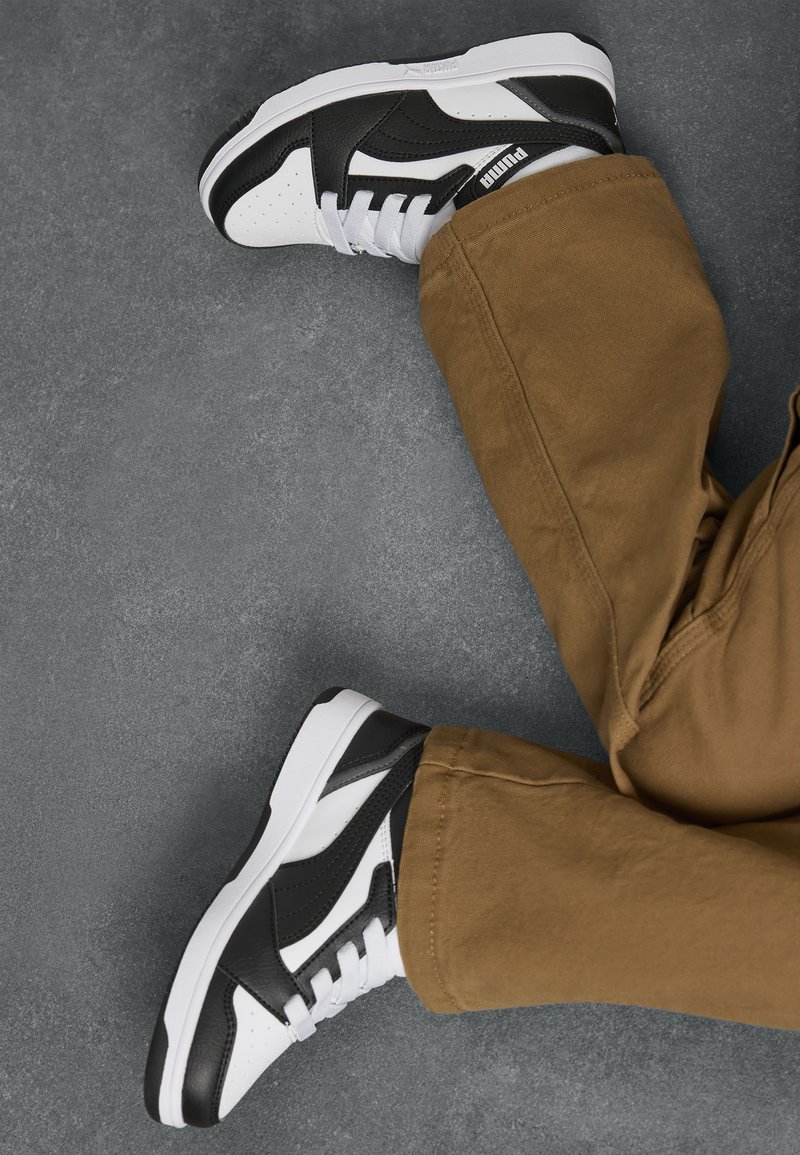 Zwart-witte sportschoenen met geperforeerde details en witte veters, gedragen met beige broek, tegen een grijze ondergrond.