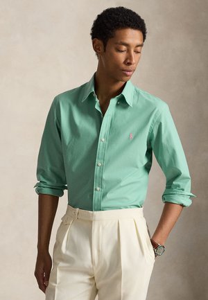 Mężczyzna o ciemnych kręconych włosach, ubrany w zieloną, zapinaną na guziki koszulę z podwiniętymi rękawami oraz kremowe plisowane spodnie, stoi na neutralnym tle.