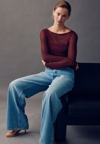 Dugotrajna vinsko crvena majica s dugim rukavima od teksturiranog pletiva u kombinaciji sa svijetloplavim trapericama širokih nogavica. Model sjedi na crnom trosjedu na sivoj pozadini.