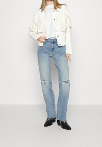 Chaqueta blanca con flecos sobre una camisa blanca, combinada con jeans claros desgastados y botines negros brillantes.