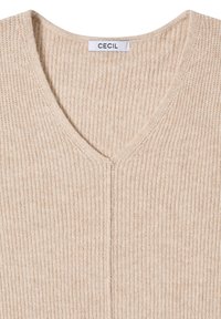 Pull en tricot beige avec un col en V, présentant une texture côtelée et une couture centrale. L'étiquette indique "CECIL" cousue dans l'encolure.