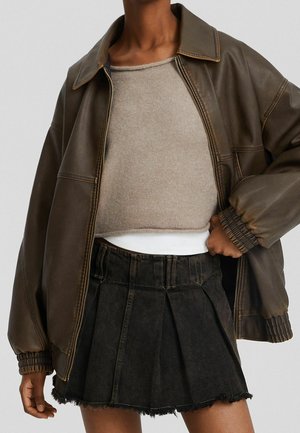 Veste en cuir marron portée sur un pull court beige et une mini-jupe plissée en denim noir à ourlet brut sur une personne debout.