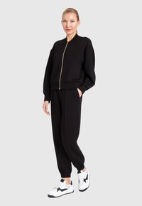 Tuta in cotone nero con giacca bomber con zip e pantaloni tapered, completata da sneakers bianche con dettagli neri.