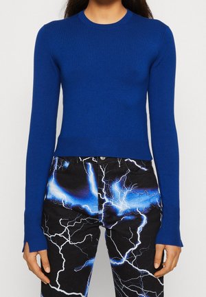 Persona con una camiseta azul ajustada de manga larga y pantalones negros con patrones de rayos en azul brillante y blanco.