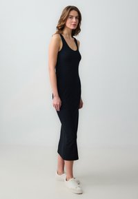 Robe noire en maille avec une silhouette ajustée, décolleté en forme de scoop et ourlet au genou. Présente un motif à carreaux subtil. Portée avec des baskets blanches.
