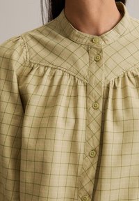 Grünes karierter Hemd mit einem hohen Kragen, gefältelten Details und Knopfverschluss. Der Stoff hat eine weiche Textur mit kontrastierenden Linien.