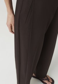Close-up of a person wearing dark brown tailored pants with a side pocket and dark brown sandals on a light background.