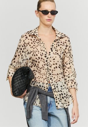 Femme portant des lunettes de soleil noires, une blouse à pois boutonnée, un jean bleu clair, un pull gris noué à la taille et tenant une pochette noire tressée.