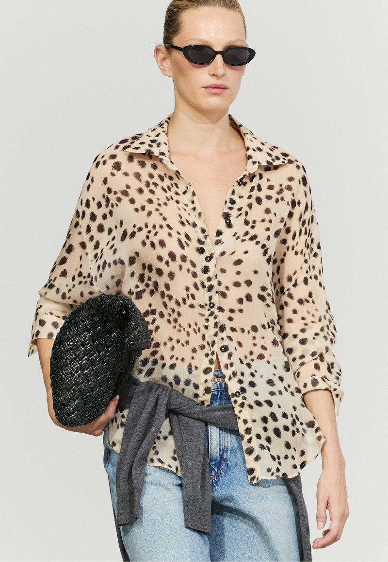 Woman in black sunglasses wears a spotted button-up blouse, light blue jeans, gray sweater tied at waist, and holds a black woven clutch.
