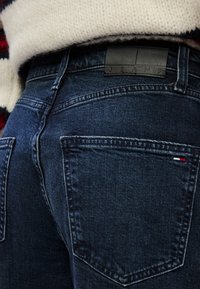 Jeans din denim albastru închis cu un finisaj texturat, cusături vizibile și o etichetă din piele neagră inscripționată "JEANS" la talie.