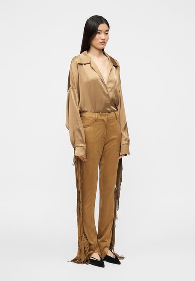 Μπεζ πουκάμισο από σατέν με μακριά μανίκια και χαμηλό γιακά, συνδυασμένο με καφέ παντελόνια με φουντάκια. Τα παντελόνια έχουν στενή γραμμή και πλαϊνές τσέπες. Μαύρα μυτερά παπούτσια.