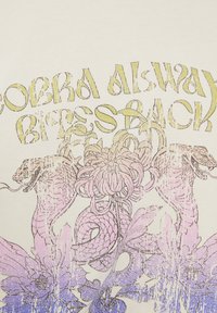 Un tejido de color crema presenta un diseño gráfico con dos serpientes entrelazadas alrededor de elementos florales en rosa y azul. El texto dice "COBRA SIEMPRE MUERDE DE VUELTA" en amarillo.
