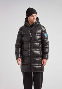 Hombre vistiendo un abrigo de plumas negro largo y brillante con bolsillos con cremallera y capucha, combinado con un gorro negro y pantalones negros, mirando hacia su izquierda.