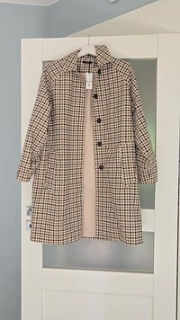 Długi, beżowo-czarny płaszcz w kratę z kołnierzem, z zapięciem na guziki i dwiema bocznymi kieszeniami, wiszący na białych drzwiach.