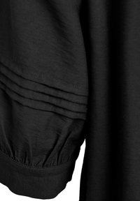 Svart plagg med en texturerad yta med pressveckdetaljer och en rynkad mudd. Tyget verkar lätt och något fallande.