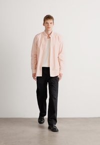 Lys rosa skjorte med knapper, hvit teksturert undertrøye, sorte bukser og sorte sko. Modellen står mot en ensfarget bakgrunn.