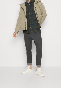 Beige softshell jakke, grønternet skjorte, grå stribede bukser og hvide sneakers. Sættet har et afslappet, lagdelt look.