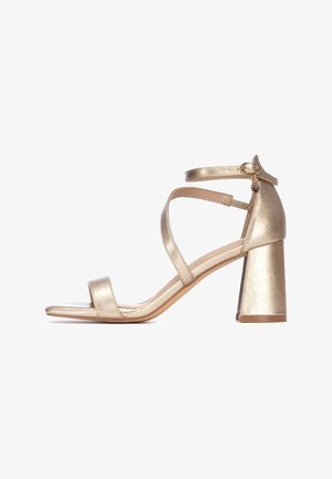 Sandalia metálica dorada con tacón grueso, correa en el tobillo y dos correas delanteras. Textura lisa y diseño de punta abierta.