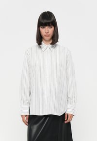 Camicia bianca con righe verticali nere, dotata di colletto classico, maniche lunghe e un taglio rilassato. Tessuto morbido al tatto.