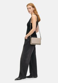Borsa a tracolla in pelle beige chiaro con una texture liscia, forma rettangolare e tracolla staccabile. Modello che indossa una canotta nera e jeans.