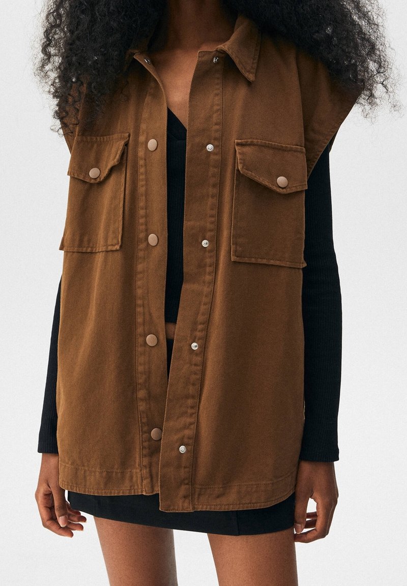 Femme portant un gilet marron sans manches boutonné avec de grandes poches à l'avant, par-dessus un haut noir à manches longues et une jupe noire.