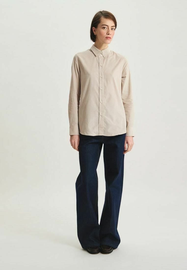 Chemise boutonnée beige clair avec col, manches longues, texture lisse ; associée à un jean foncé évasé, coupe droite, et des chaussures sombres.