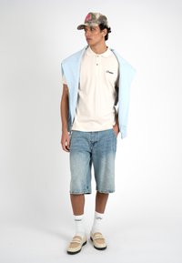 Νέος άνδρας με μπεζ πόλο, ανοιχτομπλε φούτερ περασμένο στους ώμους, σορτς από τζιν, λευκές κάλτσες, μπεζ loafers και καπέλο μπέιζμπολ με καμουφλάζ.