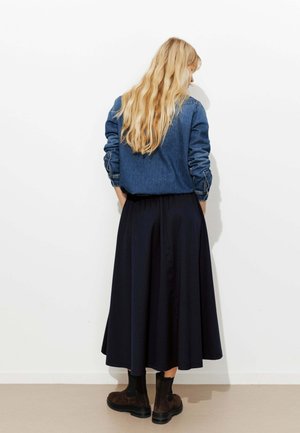 Veste en jean à manches longues, associée à une jupe plissée bleu marine longueur cheville et des bottines marron foncé, vue de dos.