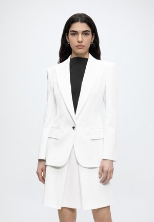 Femme aux longs cheveux foncés portant un blazer blanc ajusté et une jupe assortie par-dessus un haut noir, debout devant un fond clair uni.