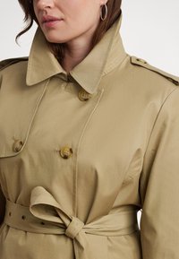 Beige színű ballonkabát nagyméretű gallérral, dupla soros gombolással és deréknál megkötött övvel. Vállpántokkal és sima textúrával rendelkezik.