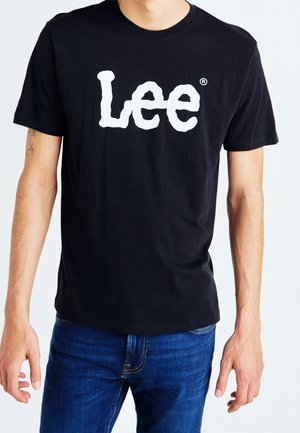 Personne portant un t-shirt noir à manches courtes avec le logo blanc « Lee » centré sur la poitrine et un jean en denim bleu sur fond blanc.