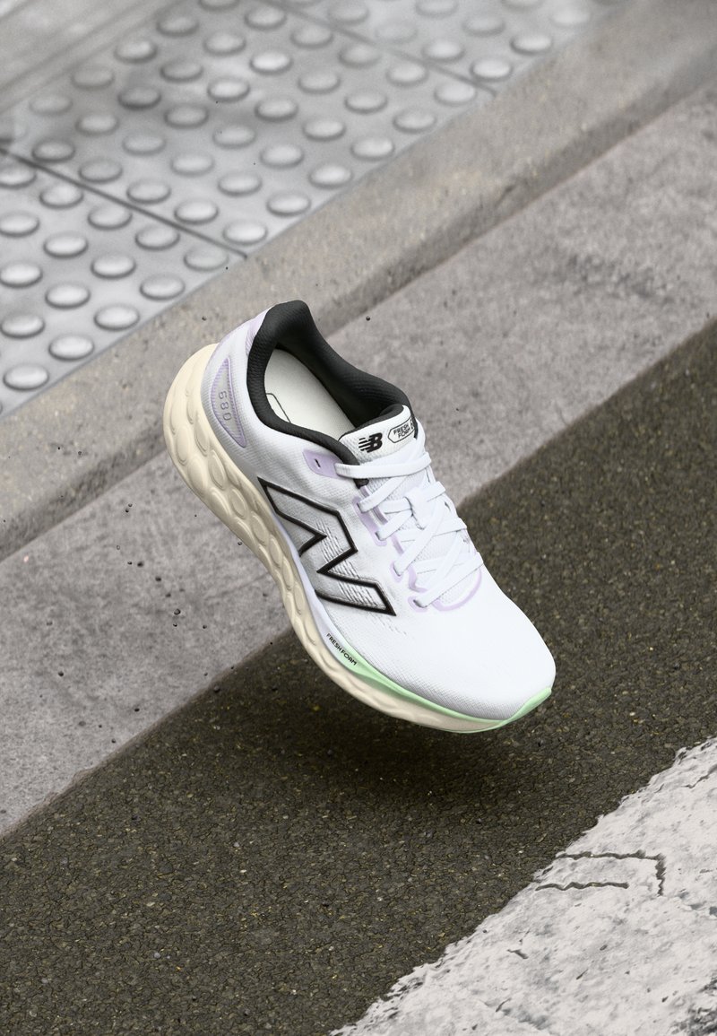 Baskets de course blanches avec des accents noirs et un logo 'N'. Elles présentent une tige texturée, un laçage et une semelle rembourrée avec une teinte verte.