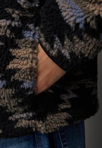 Pull en polaire avec une surface texturée en motifs noirs, beiges et bleus. Il comporte une main dans une poche latérale et un bord noir au niveau de l'encolure.