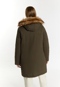 Lang olivengrøn parka med pelsforet hætte, quilted tekstur og synlige syningsdetaljer. Bagfra ses en lige kant og lommer.