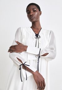 Weißes Kleid mit gerüschtem Ausschnitt und gefaltetem Stoff. Besitzt lange, durchsichtige Ärmel mit dunkelblauen Satinbändern und Ausschnitten an den Bündchen.