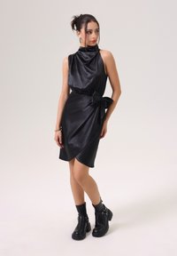 Robe en satin noir avec un col drapé, taille ceinturée avec un nœud, et ourlet asymétrique. Portée avec des bottes noires à talons épais.