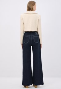 Avklippt beigt texturerat jacka med bred krage, kombinerat med mörkblå vida jeans. Har synliga sömmar och två bakfickor.