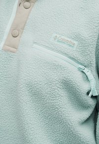 Ανοιχτό μπλε φλις πουλόβερ με μπεζ κουμπιά πατιλέτας και τσεπάκι στο σ τήθος με φερμουάρ, με ετικέτα λογότυπου της Columbia Sportswear Company.