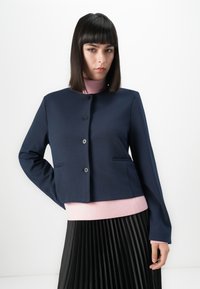 Femme aux cheveux raides et foncés portant une veste bleu marine à boutons sur un col roulé rose et une jupe plissée noire, se tenant devant un fond blanc.
