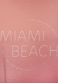 Rozā audums ar lieliem, baltas krāsas uzrakstiem "MIAMI BEACH" un mazākiem uzrakstiem: "SMILTIS UN SĀLTA ŪDENS" un "SAULE UN ZILIE DEBESS" aplī.