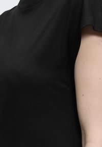 Κοντινό πλάνο του άνω κορμού ενός ατόμου που φοράει ένα απλό μαύρο μπλουζάκι με κοντά μανίκια, δείχνοντας μέρος του δεξιού χεριού.