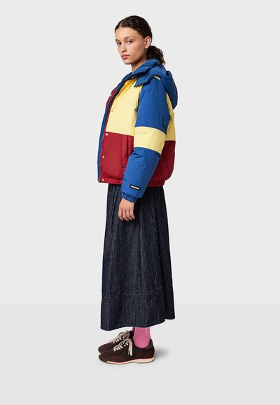 Farverig puffer-jakke med blå, gul og rød paneler. Modellen bærer en navy-stribet nederdel og mørke sko med lyserøde sokker.