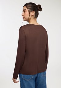 Brązowa bluzka z długim rękawem i okrągłym dekoltem, o miękkiej teksturze i luźnym kroju. Uzupełniona o niebieskie dżinsy z wysokim stanem.
