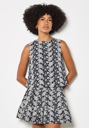 Femme aux cheveux afro portant une robe sans manches bleu marine avec une superposition de dentelle florale blanche et des boucles d'oreilles rondes en or, debout devant un fond blanc.