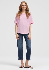 Smiling woman with long wavy hair wearing a loose pink short-sleeve top, cropped blue jeans, and black flip-flops, standing on a white background.