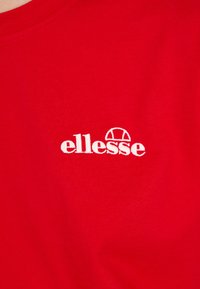 Rød bomulls t-skjorte med en hvit brodert "ellesse" logo med en halvsirkelformet detalj over teksten, som viser en jevn tekstur.